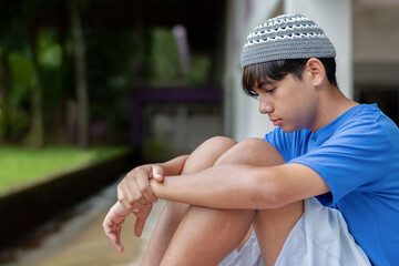 Sad teen Muslim boy sitting alone with knees up, wearing a knitted cap and blue shirt. Emotional...