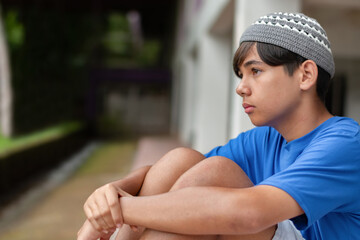 Sad teen boy sitting alone with knees up, wearing a knitted cap and blue shirt. Emotional moment of...