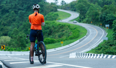 A cyclist in an orange jersey stands at the start of a winding road surrounded by lush greenery, ready to begin a challenging ride through a scenic landscape.