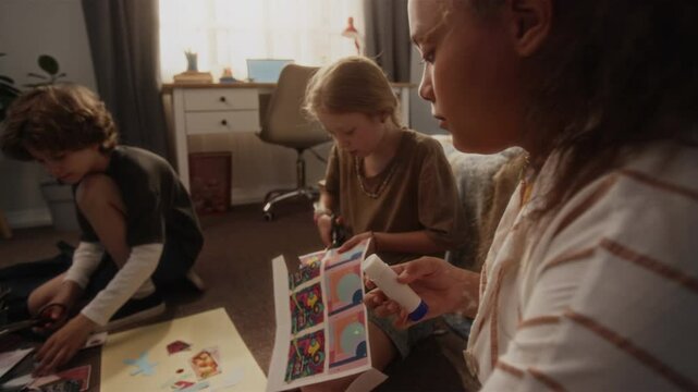 Rack focus close up shot of multiracial young girl crafting paper collage using glue stick while friends cutting out pictures decorating cut and paste artwork together on floor in bedroom