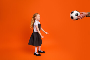 Happy young schoolgirl in uniform learning to catch a soccer ball against a vibrant orange...
