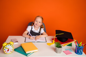 Adorable schoolgirl writing in a notebook at a desk surrounded by school supplies on an orange...