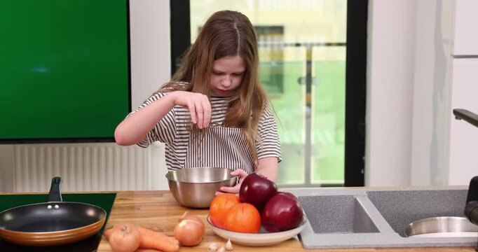 Focused girl plays pouring dry ingredients into stainless bowl in kitchen. Schoolgirl with loose hair learns to cook standing at wooden counter