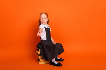 Adorable schoolgirl in uniform sitting on stacked books holding hand out against orange background