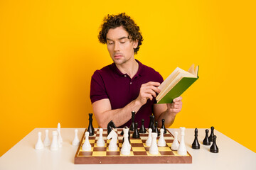 Young man reading a book while analyzing chessboard pieces on a bright yellow background, dressed...