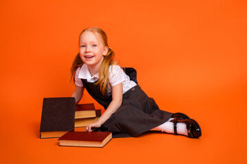 Young schoolgirl in uniform sitting with books against a bright orange background, showcasing...