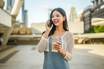 Young Asian successful business woman standing in big city talking on mobile phone and savoring her coffee , Smiling and enjoying a casual conversation, bright daylight, Modern Urban Lifestyle