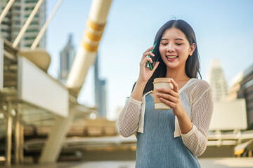 Young Asian successful business woman standing in big city talking on mobile phone and savoring her coffee , Smiling and enjoying a casual conversation, bright daylight, Modern Urban Lifestyle