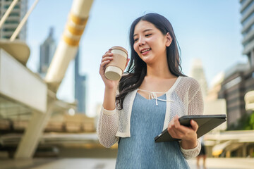 A joyful asian young business woman takes a moment to relax, savoring her coffee while engaging with a tablet. The lively city backdrop adds energy to her break on a bright day.