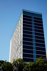 Obraz premium Tall modern office tower with glass facade reflecting clear blue sky. Minimalist architecture emphasizes vertical lines, symmetry, and urban elegance in city landscape.