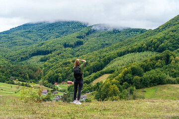 Back view of travel photographer and vlogger shooting a panoramic view of forested mountains and a village below.Woman hiker with a backpack enjoying the stunning view. Concept of freedom, adventure