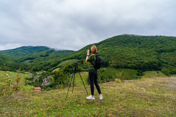Woman photographer with a tripod capturing a stunning mountain landscape and a village below. Female traveler taking photos of a scenic mountain valley on a cloudy day in the Carpathians