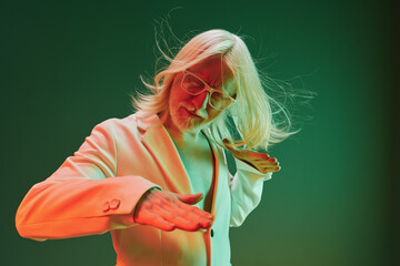 Stylish older man with long hair and glasses performing a dance gesture against a gradient green...