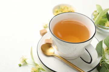 Tasty linden tea in cup, spoon, sugar cubes, leaves and flowers on white table, closeup. Space for text