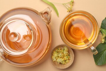 Tasty linden tea in cup, teapot, leaves and flowers on beige table, flat lay