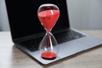 Deadline. Hourglass with flowing sand and laptop on wooden table indoors, closeup