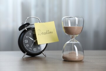 Sticky note with word Deadline, hourglass and alarm clock on wooden table indoors
