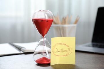 Sticky note with word Deadline, hourglass, stationery and laptop on wooden table indoors, closeup