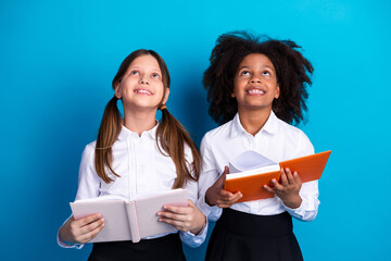 Two cheerful schoolgirls in uniforms smiling while holding book against a vivid blue background,...