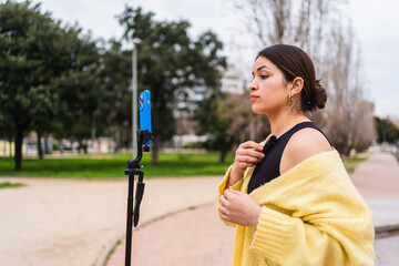 Young woman attaching her lavalier microphone while preparing to record a video with her smartphone on a tripod in a park