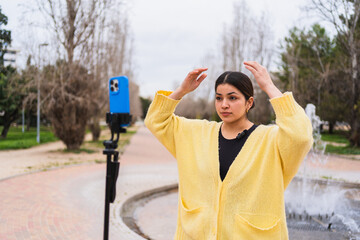 Young woman adjusting her hair while recording a video blog in a park, using a smartphone on a tripod with a lavalier microphone