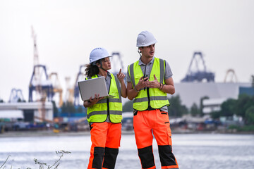 Two engineers wearing safety vests and helmets inspect port operations near the waterfront. They...