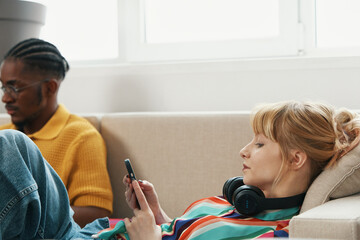 Young adult woman working from home with headphones resting, while male colleague in background...