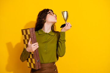 Young woman holding a chessboard and golden trophy on a yellow background, celebrating success in a...