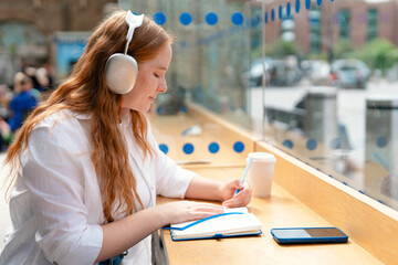 Young woman with headphones writes in notebook at urban cafe during daylight hours surrounded by...