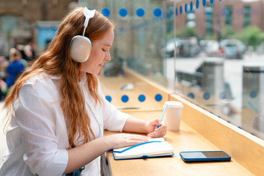 Young woman with headphones writes in notebook at urban cafe during daylight hours surrounded by city life