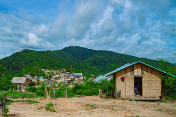 Fototapeta premium A traditional bamboo house with roof stands in the foreground, overlooking a sprawling rural village nestled within a valley of lush green mountains. 