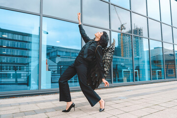 Woman with angel wings poses creatively in front of modern building during daytime with clear sky...