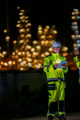Fototapeta premium Female engineer in high visibility jacket uses a radio while analyzing data on a laptop during a night inspection at an industrial site, with blurred lights in the background.