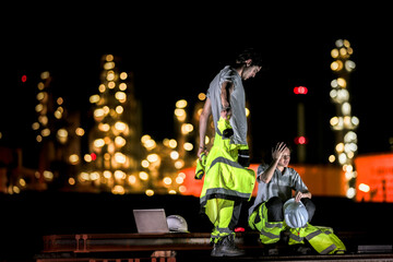 Two industrial workers take a break during the night shift at a construction site, sitting on steel...