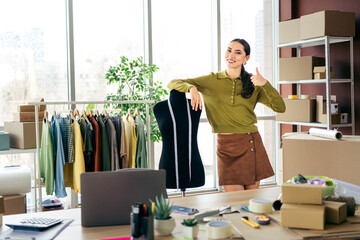 Young female entrepreneur standing confidently in a fashion atelier surrounded by clothing and...