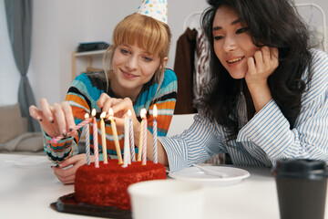Two young women coworkers lighting birthday candles on cake, socializing and celebrating in workplace, representing Gen Z friends bonding during work break.