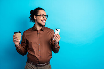 Confident man in brown shirt holding smartphone and coffee cup, on blue background, embodies modern...