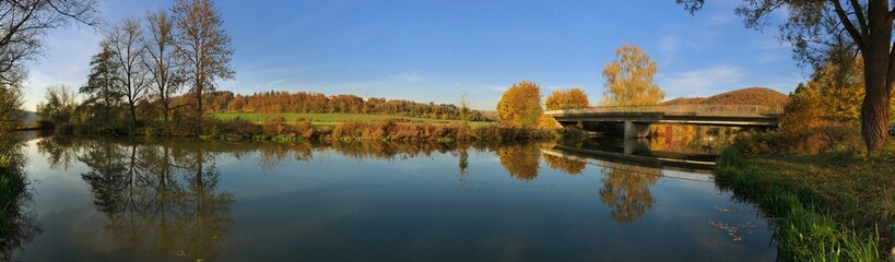 Obraz premium Wide panoramic view of an autumn river with reflections and a bridge.