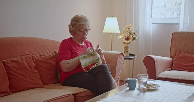 Senior woman knitting on the sofa in her living room. She is enjoying her retirement and hobbies.