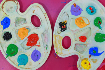 Bright acrylic paint blobs arranged on two plastic palettes over pink table.	Colorful thick acrylic...