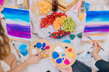 Two canvases showing vivid gradient landscapes are being painted during an outdoor art picnic with...