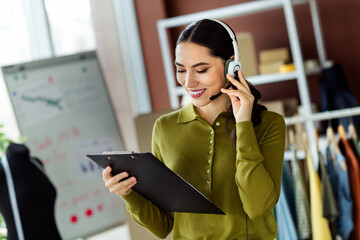 Confident businesswoman at her fashion workshop wearing a headset and holding a clipboard, engaged...
