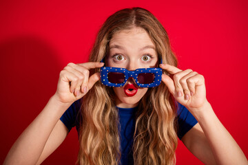 Cheerful young girl with stylish sunglasses making a surprised expression against a vibrant red...