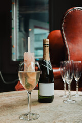 Wine glass with grapefruit popsicle champagne cocktail and mint sprig, set on rustic wooden table with champagne bottle, flutes, red leather wingback chair and decorative lights