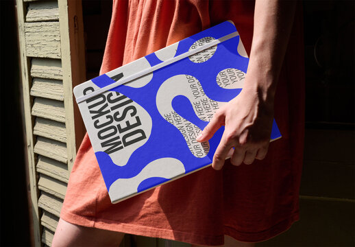 Woman holds a clean notebook mockup, showcasing its cover against a warm, sunlit backdrop. Perfect for creative projects. PSD mockup