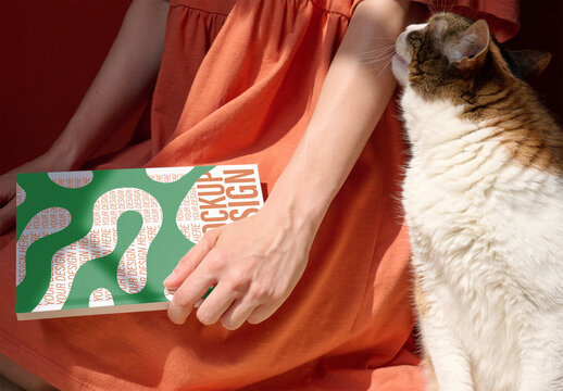 Female hand holds a blank notebook while a cat curiously approaches in a warm indoor atmosphere. PSD mockup.