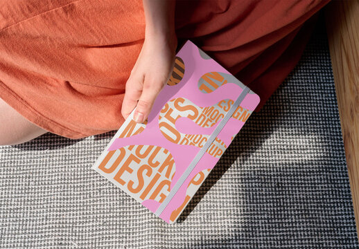 Woman sits on a patterned surface, holding a clean, modern notebook. PSD mockup.