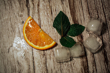 A vibrant orange slice, fresh mint leaves, and ice cubes artfully arranged on a rustic wooden...