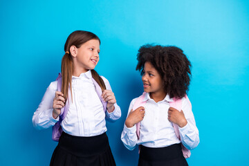 Two cheerful schoolgirls in uniforms with backpacks sharing a friendly smile against a blue...