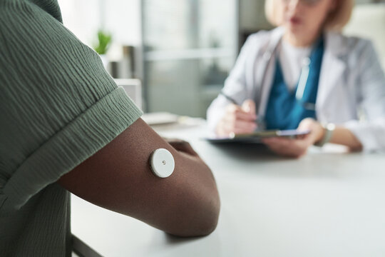 Black young adult man wearing glucose monitor on upper arm consulting with Caucasian middle aged female doctor in medical office, doctor writing notes during diabetes management appointment